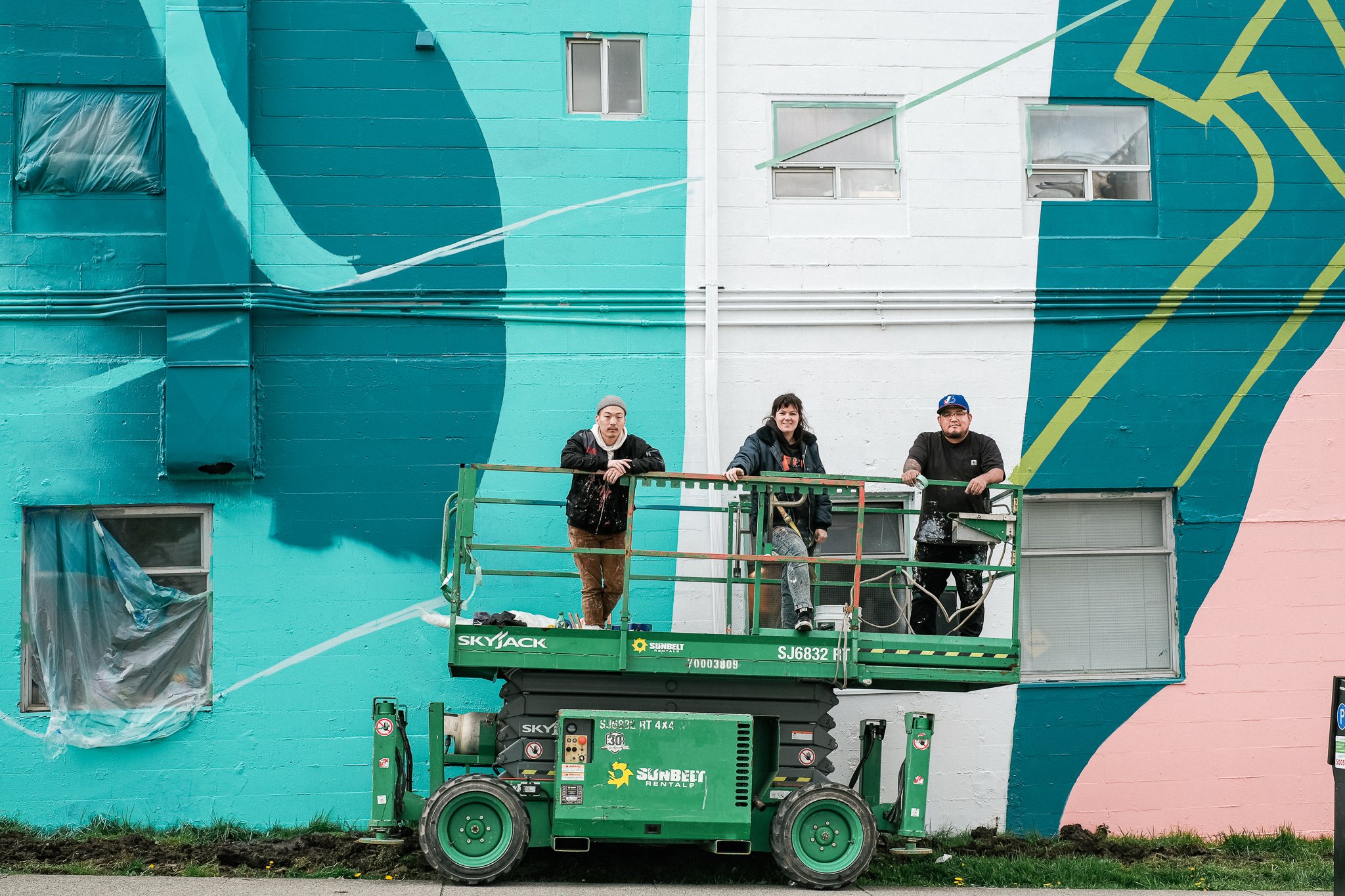 Artists, Joon, Fiona and KC stood on green scissor lift in front of their in progress mural 